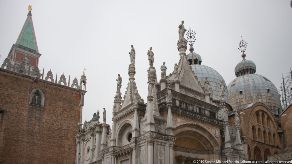 Gothic pinnacles and statues rise before lead-clad domes and brick architecture under an overcast Venetian sky.