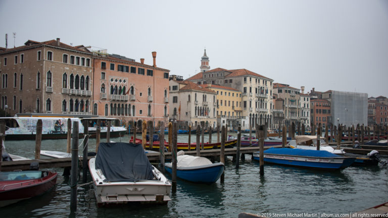 Small boats moored among timber posts on the Grand Canal before a long row of colorful Venetian palazzi.