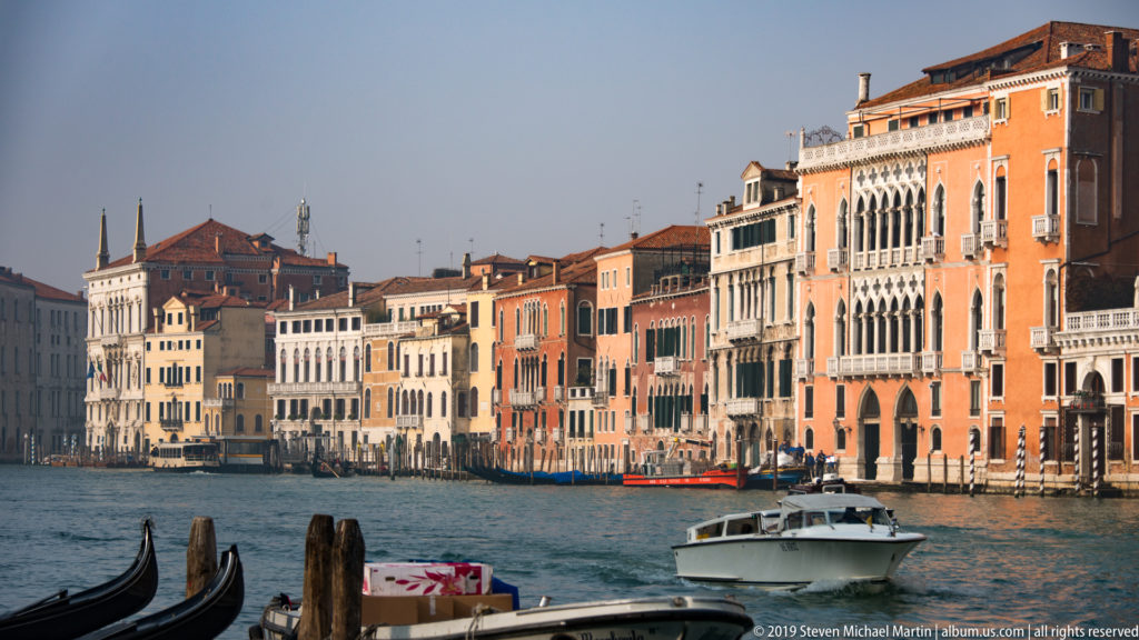 Sunlit Grand Canal lined with historic palazzi, with a motorboat crossing and gondola equipment in the foreground.