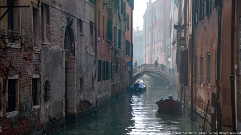 Narrow canal between weathered buildings with two small boats and a low arched footbridge in the distance.