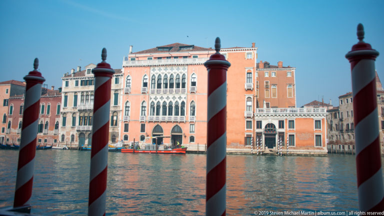 Venetian Gothic palace across the Grand Canal framed by red-and-white striped mooring poles in the foreground.