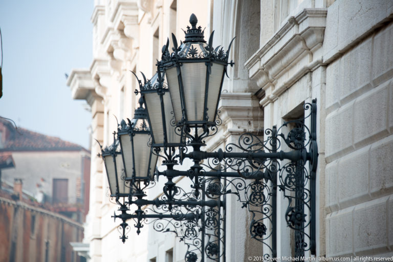 Row of ornate black wrought-iron lanterns projecting from a pale stone Venetian façade.