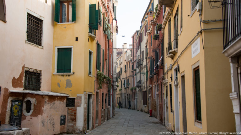 Narrow Venetian alley lined with yellow and red façades, shuttered windows, exposed brick, and a distant pedestrian.
