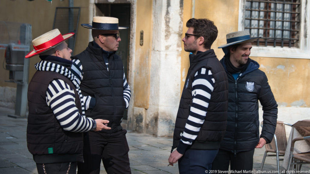 Four gondoliers in striped shirts, dark vests, and brimmed hats converse beside a weathered Venetian building.