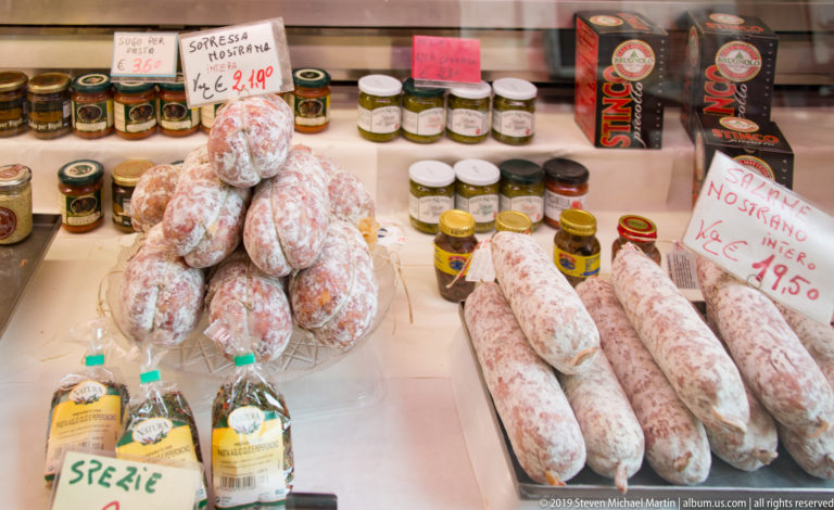 White-dusted cured salami stacked beside jars in a shop window with a handwritten 'Salame Nostrano Intero' price sign.