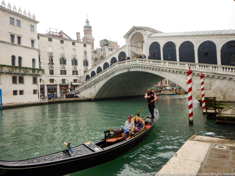 Passenger gondola crossing green canal water before the Rialto Bridge, with a standing gondolier and red-and-white mooring poles.