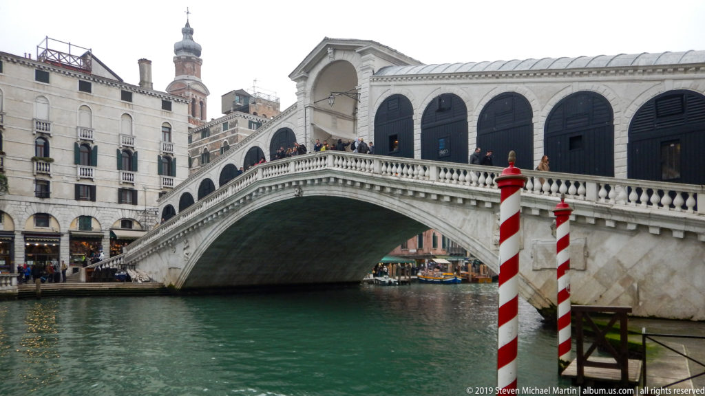 Three-quarter view of the Rialto Bridge with its broad stone arch, shop openings, balustrades, and red-and-white mooring poles.