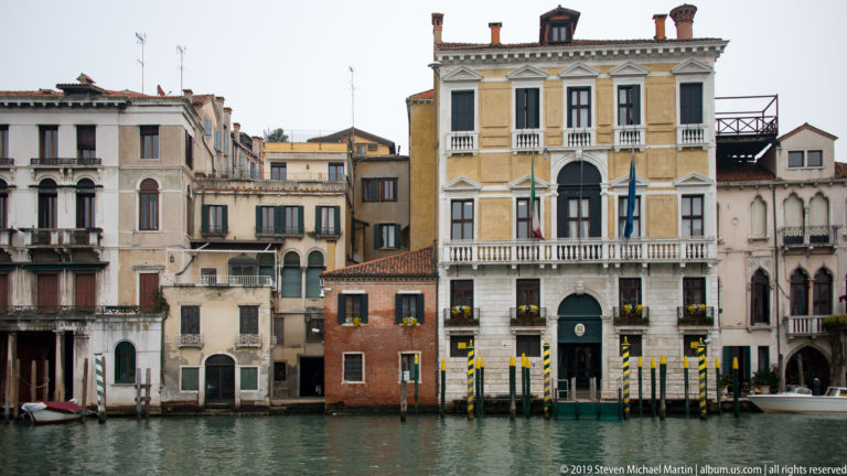 Adjoining Grand Canal palazzi centered on a cream façade with balconies and flags, fronted by green-and-yellow striped poles.
