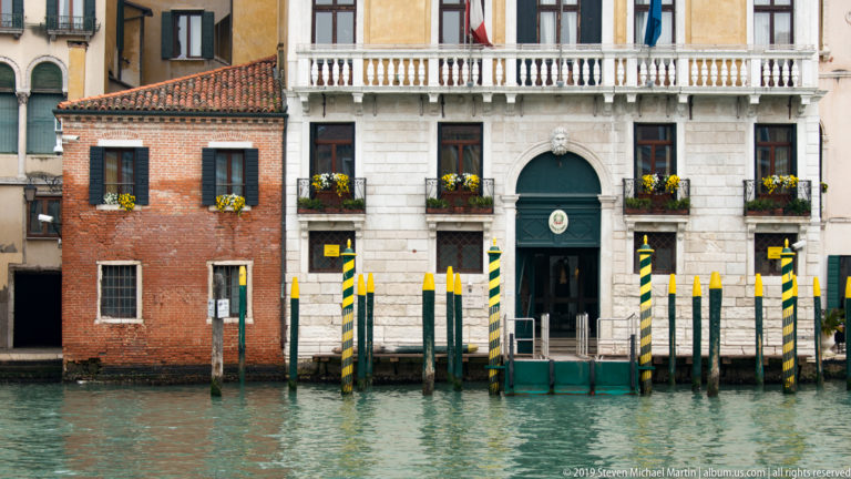 Pale canal-side palazzo with central arched doorway, balcony, flags, flower boxes, and green-and-yellow striped mooring poles.