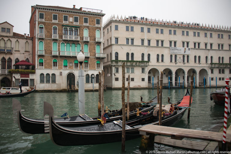 Black gondolas moored among timber posts on the Grand Canal, with pale and brick Venetian palazzi across the water.