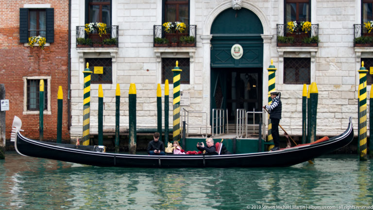 Passenger gondola beside green-and-yellow striped mooring poles, with a gondolier standing at the stern before a pale arched doorway.