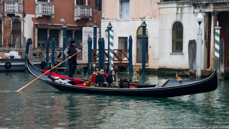 Black gondola carrying three passengers as a striped-shirt gondolier rows past canal-side buildings and mooring poles.