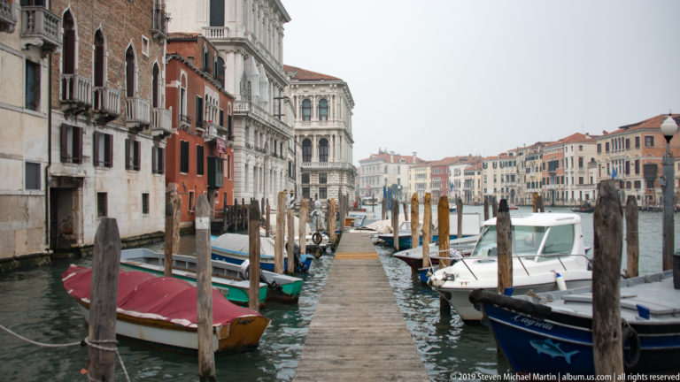 Wooden landing extending between moored small boats and timber posts toward a canal lined with Venetian buildings.