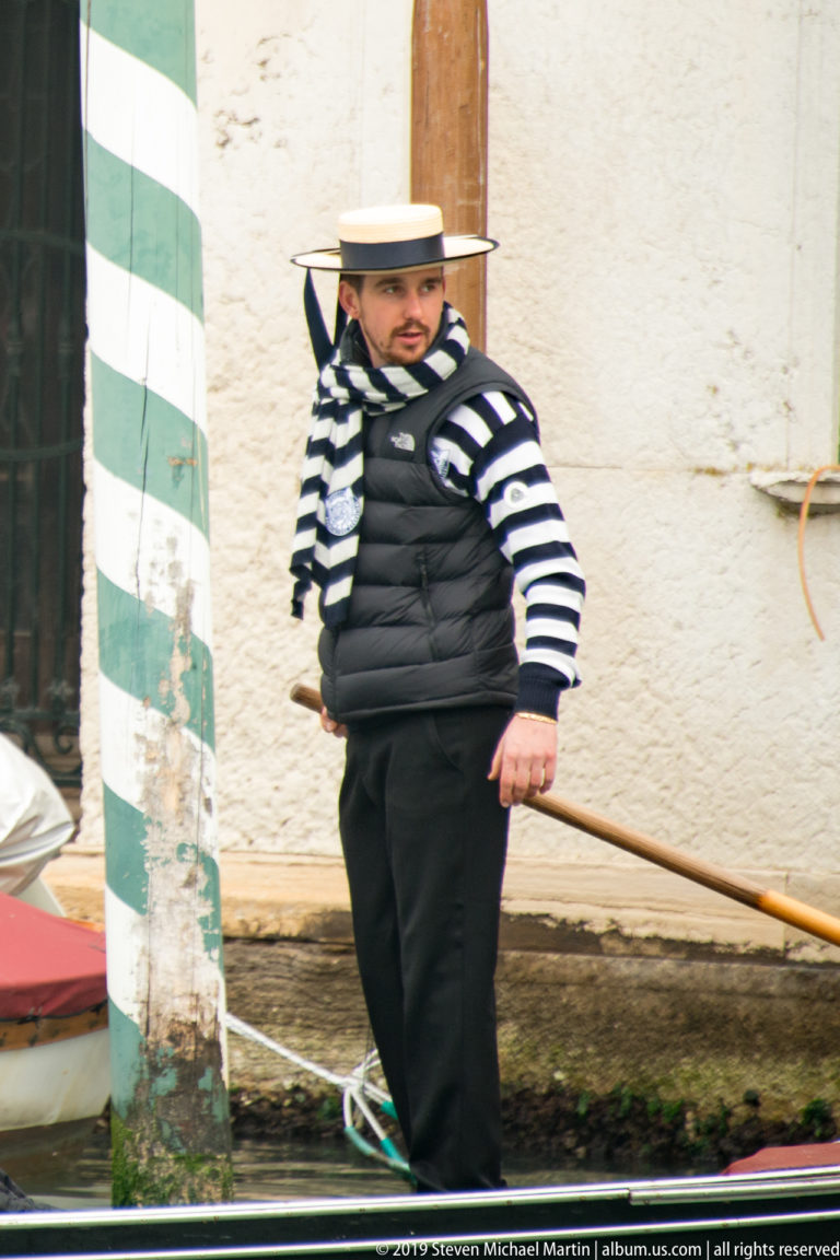Gondolier in straw hat, striped sweater, dark vest, and scarf standing beside a green-and-white mooring pole.