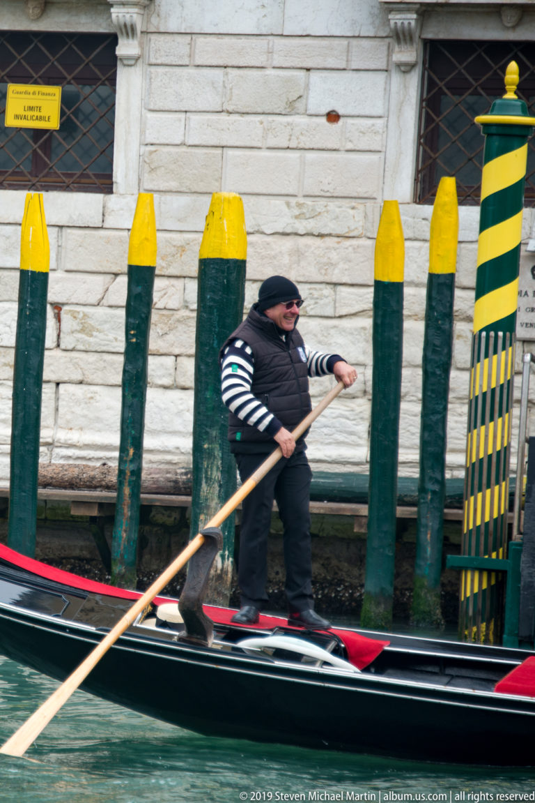 Gondolier in striped shirt and dark vest rowing a black gondola beside green-and-yellow mooring poles.