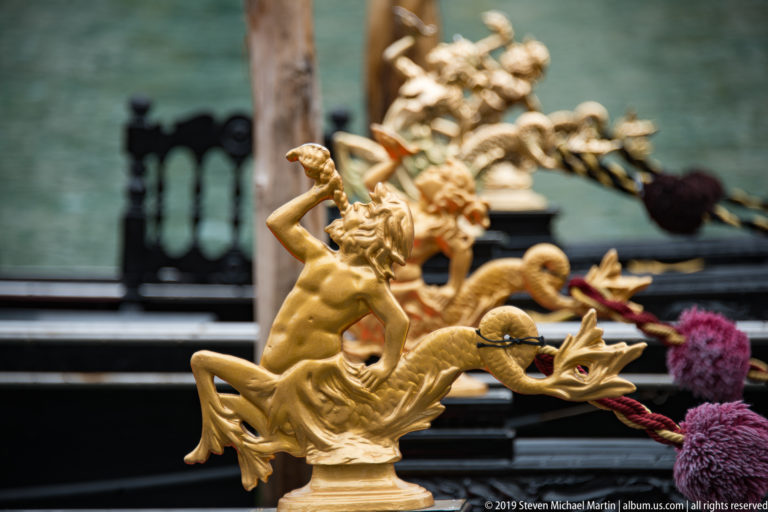 Gilded gondola ornament with a small human figure and curling animal-like forms against dark boat fittings and canal water.
