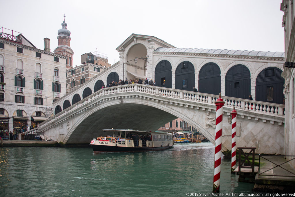 Rialto Bridge over the Grand Canal with a vaporetto passing beneath and red-and-white mooring poles at right.