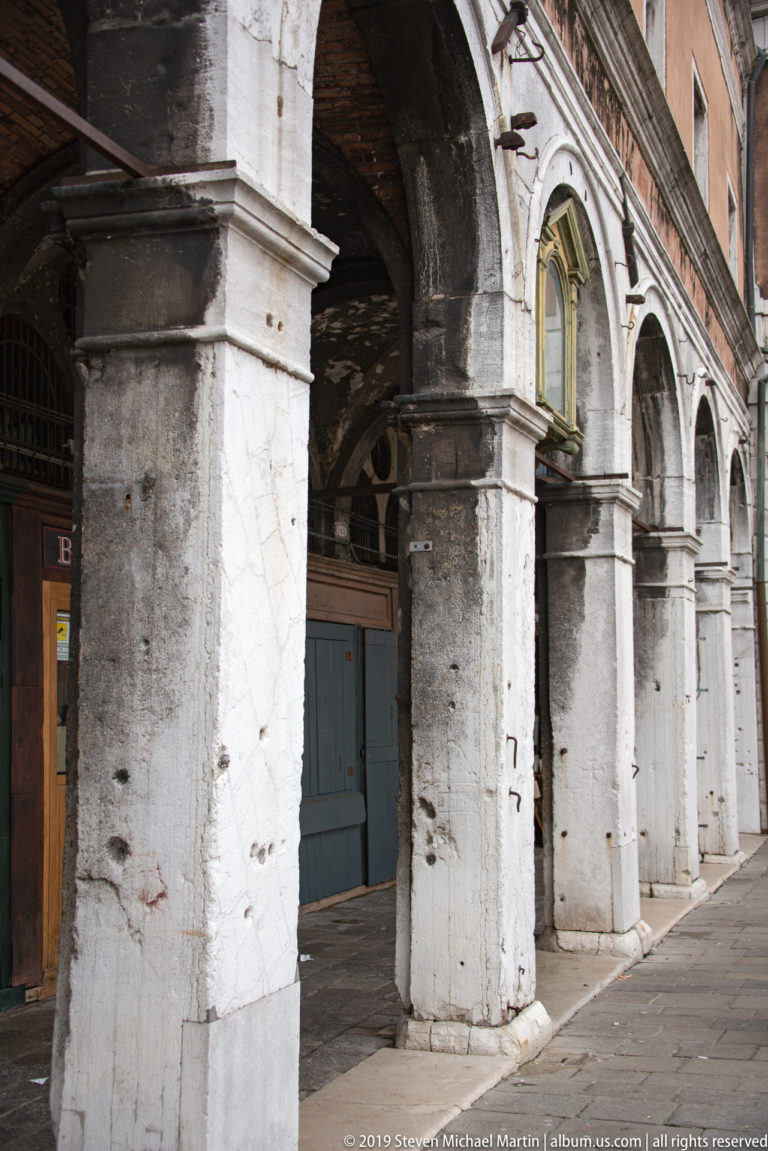 Weathered stone arcade with massive piers, round arches, recessed doors, and iron grilles along a covered walkway.