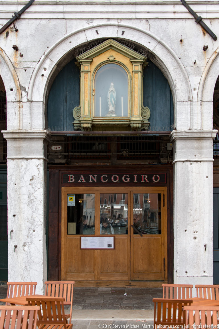 Banco Giro wooden storefront beneath a large stone arch, with a framed devotional image above and benches in front.