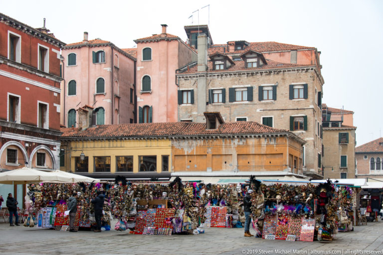 Outdoor souvenir stalls crowded with colorful merchandise beneath old Venetian residential façades.
