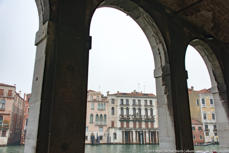Large stone market arches in deep shade framing pale canal-side palazzi across the water.