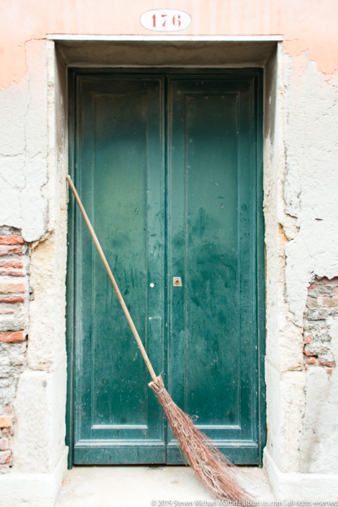 Weathered green double door numbered 176 with a twig broom leaning diagonally across patched stucco and exposed brick.