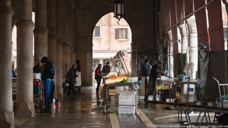 Fish vendors and illuminated counters beneath a stone market arcade with massive columns, arches, wet paving, and crates.