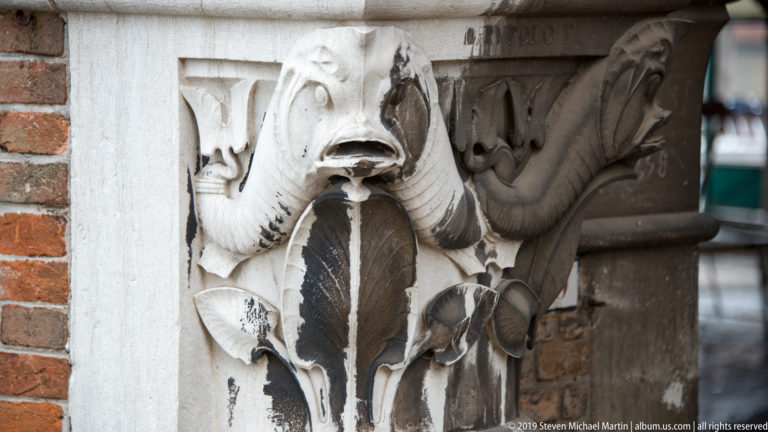 Weathered pale stone capital carved with a grotesque face, curling foliage, and elongated animal forms beside brickwork.