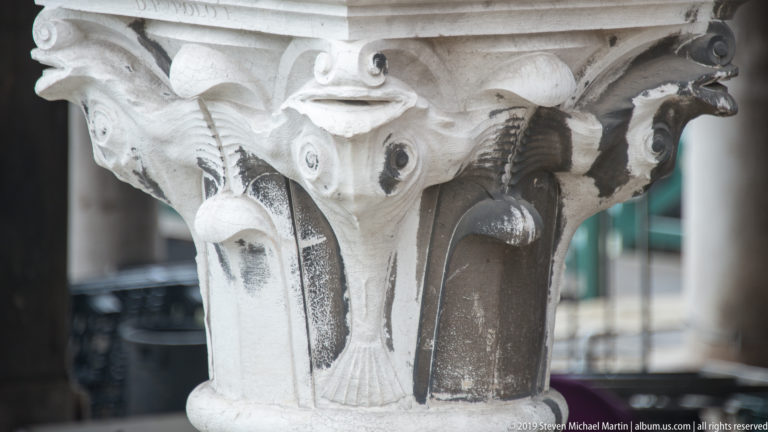 Close-up of a pale carved stone capital with curling foliage, scaled marine forms, and projecting animal heads.