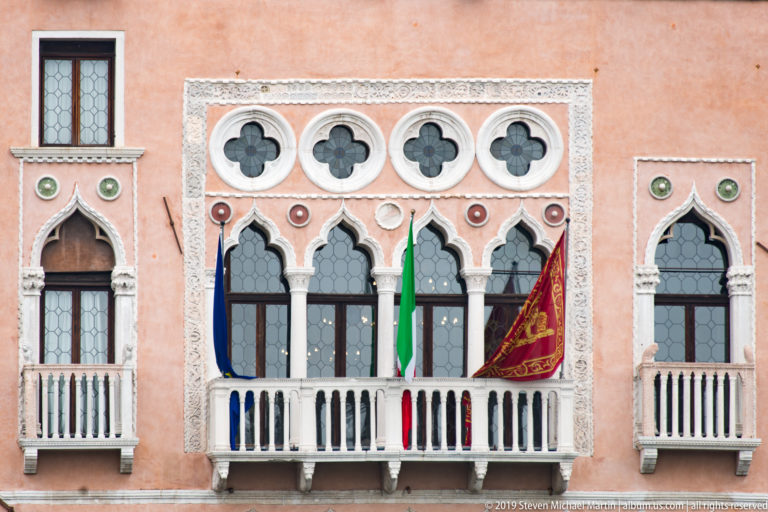 Salmon façade with pointed Gothic windows, quatrefoils, white carved trim, balconies, and hanging flags.