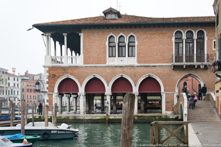 Red-brick canal-side building with pointed ground-floor arches, Gothic windows, a small loggia, and an adjoining footbridge.