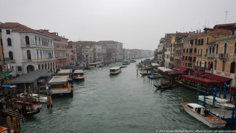 Elevated Grand Canal panorama with vaporetti, gondolas, motorboats, awnings, and historic palazzi lining both banks.