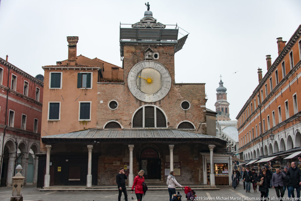 San Giacomo di Rialto with its large circular clock above a columned portico, framed by arcaded buildings and pedestrians.