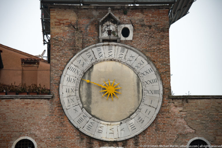 Large twenty-four-hour clock with Roman numerals and a gilded sunburst hand mounted on the brick façade of San Giacomo di Rialto.