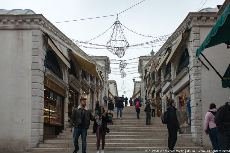 Pedestrians on the stepped, shop-lined walkway of the Rialto Bridge beneath suspended decorative lights.