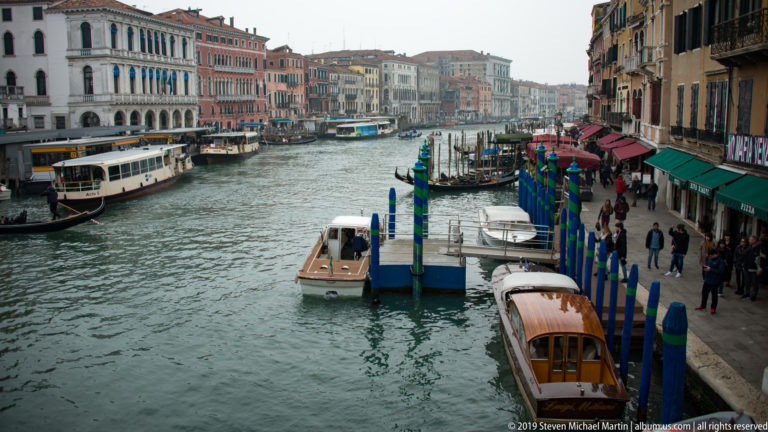 Elevated Grand Canal view with small boats, striped mooring poles, awnings, pedestrians, and colorful palazzi.