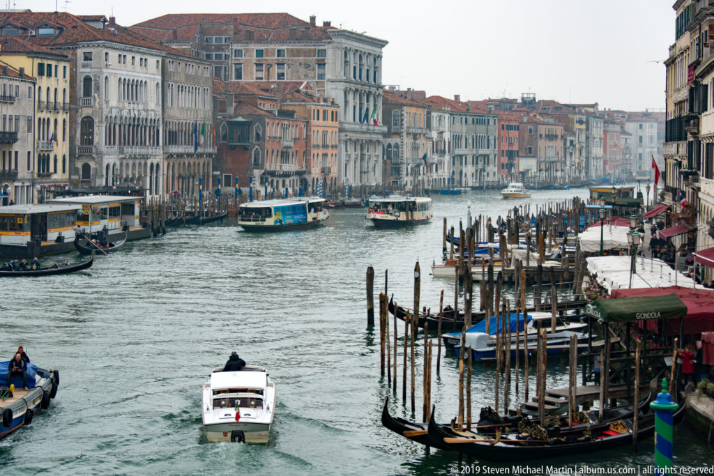 Grand Canal with a white motorboat, vaporetti, gondolas, dense mooring posts, and palazzi viewed from above.