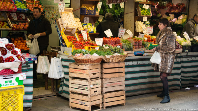 Shopper with a white bag standing at a crowded Rialto produce stall while vendors work behind crates of fruit and vegetables.