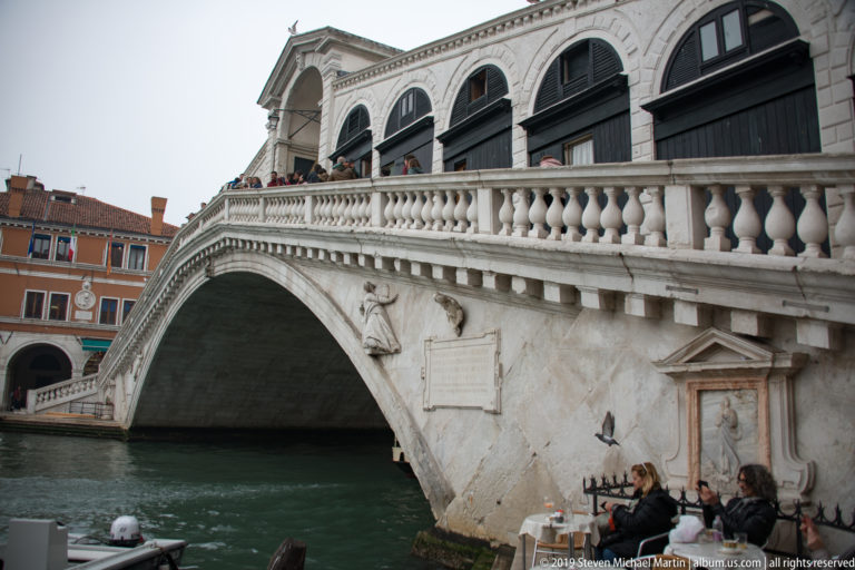 Rialto Bridge over the Grand Canal with seated musicians performing at the waterfront in the lower right.