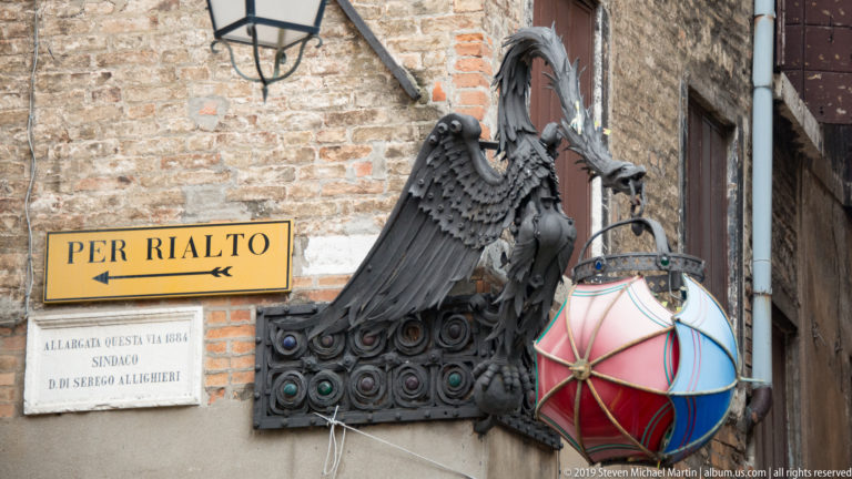Yellow 'PER RIALTO' arrow sign on a brick wall beside ornate metalwork and a colorful hanging lamp.