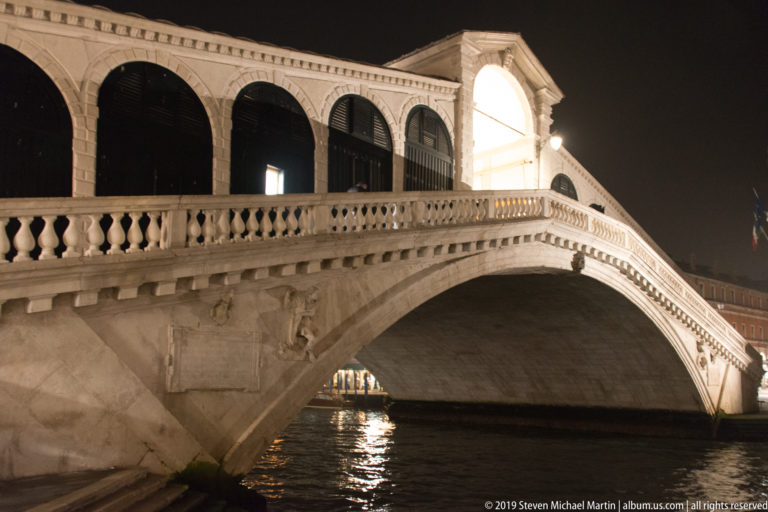 Night detail of the Rialto Bridge showing its stone arch, carved panel, balustrade, and dark shop openings.