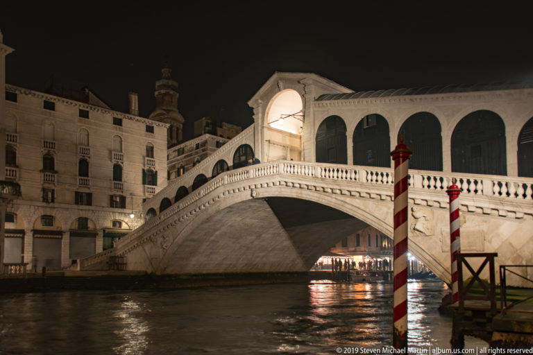 Illuminated Rialto Bridge at night above dark canal water, with two red-and-white mooring poles at right.