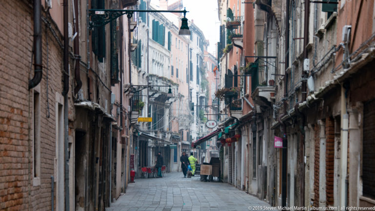 Narrow Venetian shopping street lined with brick and stucco façades, shutters, signs, balconies, and a few pedestrians.
