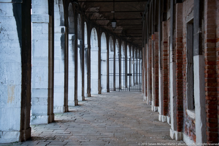 Covered Rialto walkway with red-brick piers, pale stone columns, worn paving, hanging lamps, and canal light at left.
