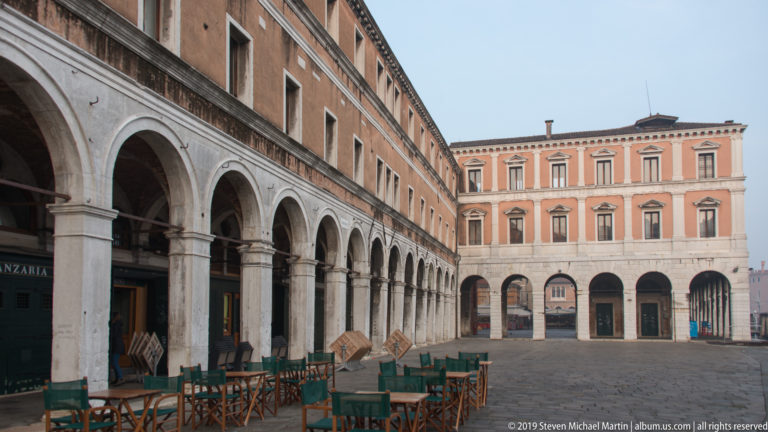 Open paved Rialto market square bordered by long arcades, with café tables beneath the arches.