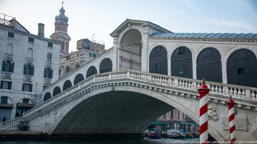 Rialto Bridge spanning the Grand Canal with its stone arch, balustrades, shop openings, and red-and-white mooring poles.