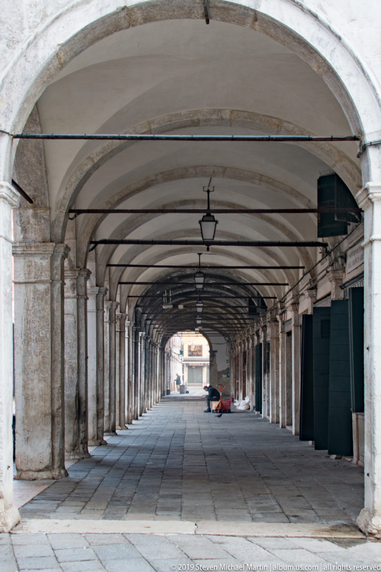 Long empty portico with repeated stone columns, shallow vaults, hanging lanterns, and worn paving in the Rialto district.