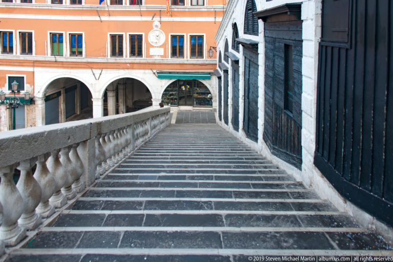 Long stone stairway on the Rialto Bridge descending between a balustrade and shuttered shops toward an orange building.