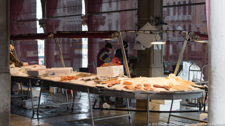 Seafood market counter with fish and shellfish under task lights between stone columns on wet paving.
