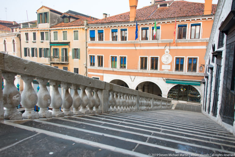 Stone steps descend along the Rialto Bridge balustrade toward an orange arcaded building with flags and shuttered windows.